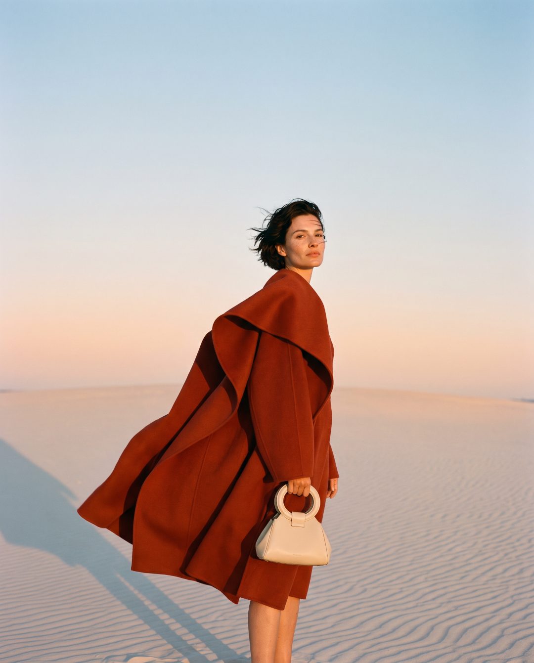 A model in a rust-red coat on a sand dune at golden hour